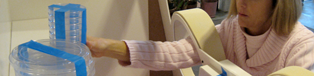 Close up of a woman in a pink sweater reaching her arms through a protective laboratory enclosure to reach for a stack of clear disks taped together with blue painters tape, approximating the size, shape, and weight of a product design still in prototype.