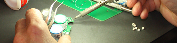 Close-up of a pair of hands soldering a circuit board on a well-lit black desk.