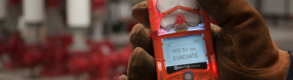 A gloved hand hods the Ventis Pro personal gas monitor while a field site is visible in the background