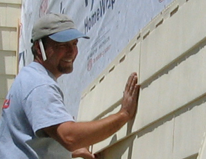 A construction worked is shown attaching cream-colored cedar shake siding to a house.