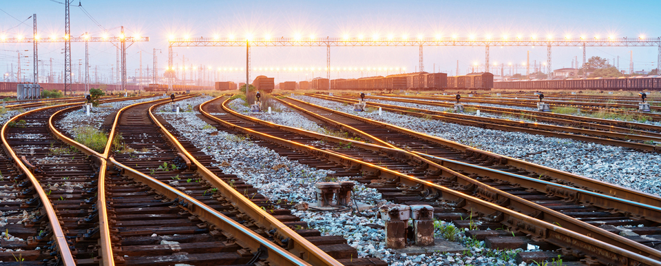 Several sets of railroad tracks merge and cross and disappear into the background, which shows a train yard in the far background that is lit by a row of bright lights.