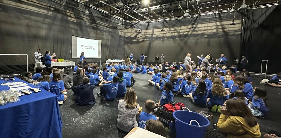 About 50 kids wearing blue Design Lives Here t-shirts are sitting in a large warehouse-like room and are in front of a projector screen showing a time limit for the current activity. Various adults are walking through the groups helping to explain things.