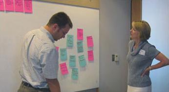 Two people stand in front of a white board with several sticky notes attached to it and discuss those ideas.