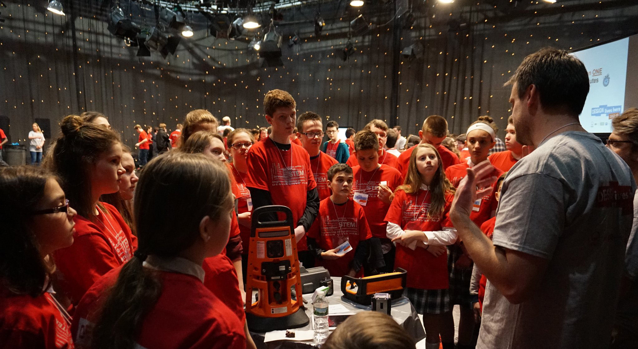 A Daedalus employee is standing at a table surrounded by about 20 kids attending Design Live Here. The Daedalus employee is demonstrating products developed by Daedalus, including an area gas monitor and a mining remote control.